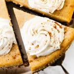 A close-up of a Pumpkin Spice Cheesecake with a slice being lifted out. The cheesecake is topped with swirls of whipped cream and a light sprinkle of cinnamon.