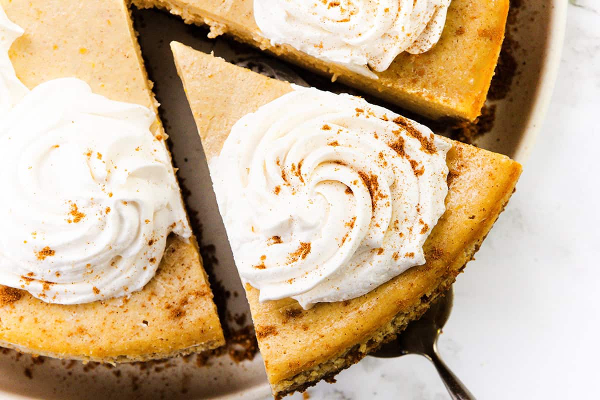 A close-up of a Pumpkin Spice Cheesecake with a slice being lifted out. The cheesecake is topped with swirls of whipped cream and a light sprinkle of cinnamon.