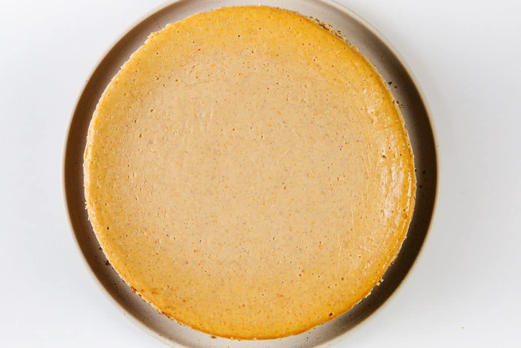 A plain, round, golden-brown Pumpkin Spice Cheesecake sits centered on a neutral-colored plate against a white background. The cheesecake has a smooth, glossy surface with no toppings or decorations.