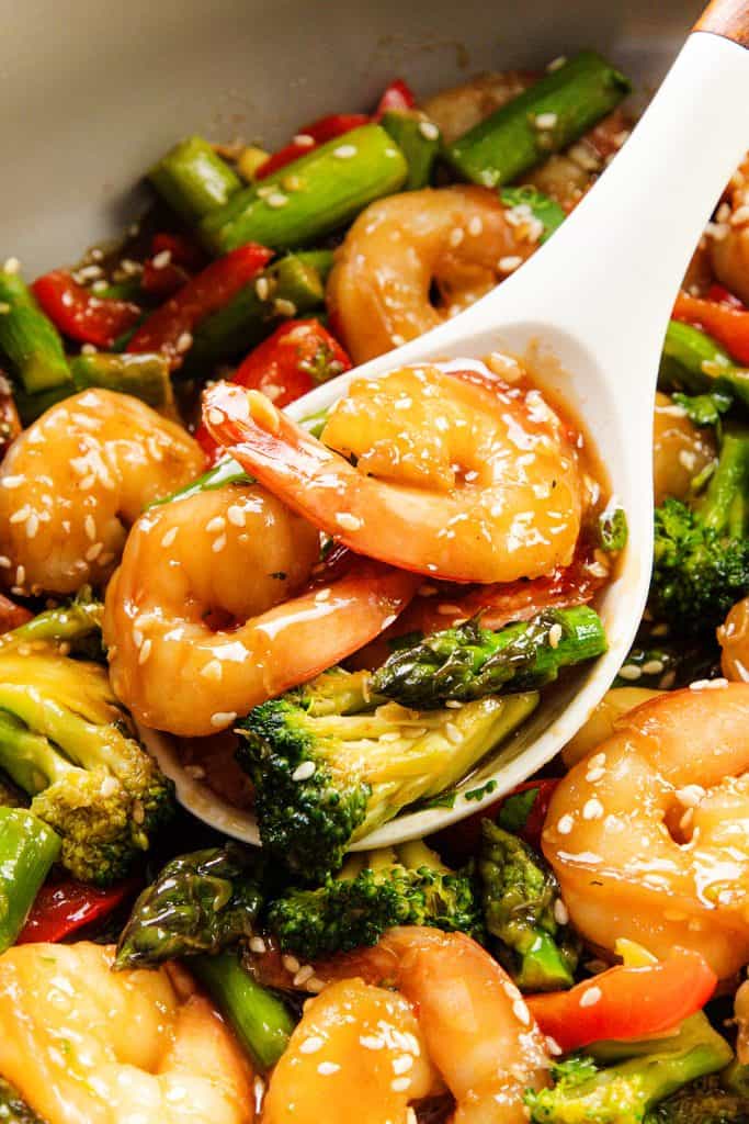 A close-up of a white spoon scooping vibrant shrimp, broccoli, asparagus, and red bell peppers coated in a glossy sauce and topped with sesame seeds—an Easy Shrimp Stir that's colorful and full of flavor.