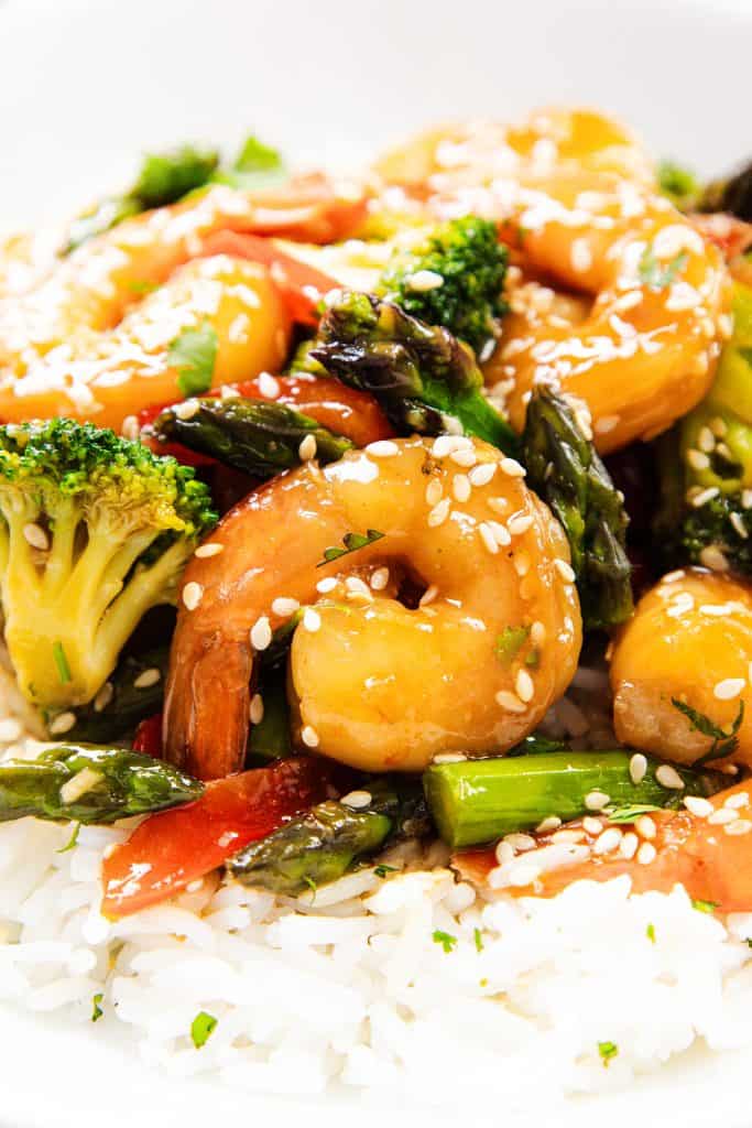 A close-up of shrimp stir-fry with broccoli, asparagus, and red bell pepper served over white rice, topped with white sesame seeds.