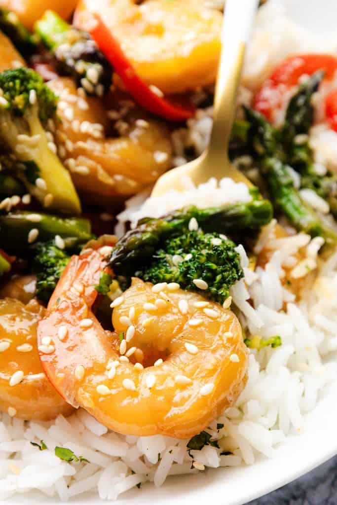 Close-up of an Easy Shrimp Stir plate featuring shrimp, broccoli, asparagus, red bell peppers, and sesame seeds over white rice, with a fork partially visible.