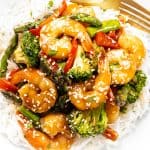 A plate of white rice topped with shrimp stir fry—broccoli, asparagus, and red bell peppers—garnished with sesame seeds and herbs. A gold fork rests on the side, perfect for a quick shrimp dinner.