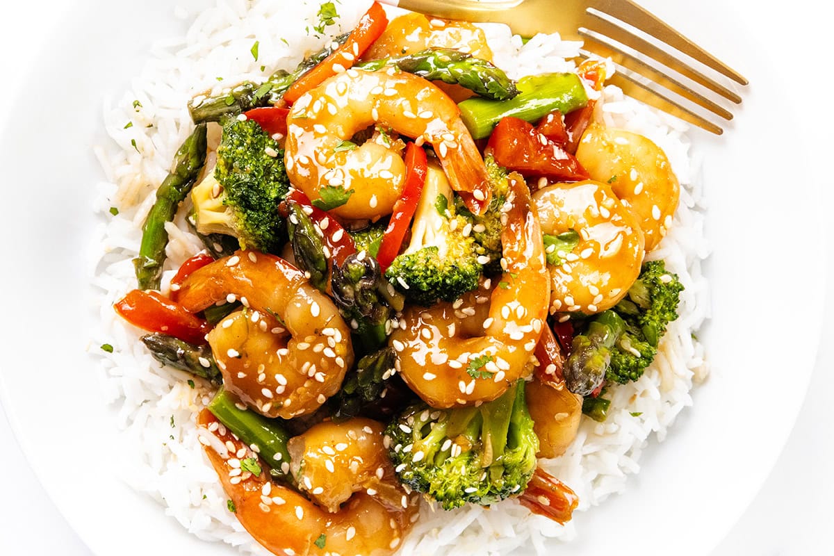A plate of white rice topped with shrimp stir fry—broccoli, asparagus, and red bell peppers—garnished with sesame seeds and herbs. A gold fork rests on the side, perfect for a quick shrimp dinner.