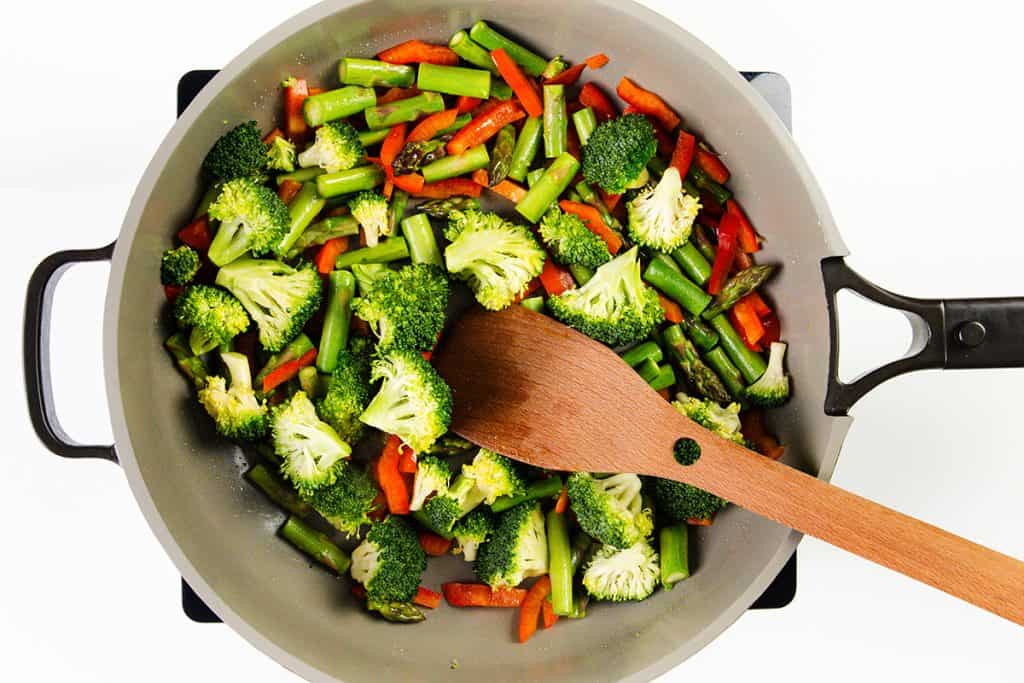 A frying pan on a stovetop filled with chopped broccoli, red bell pepper, and asparagus being stirred with a wooden spatula—a vibrant base for a quick dinner or an easy shrimp recipe like Shrimp Stir Fry.