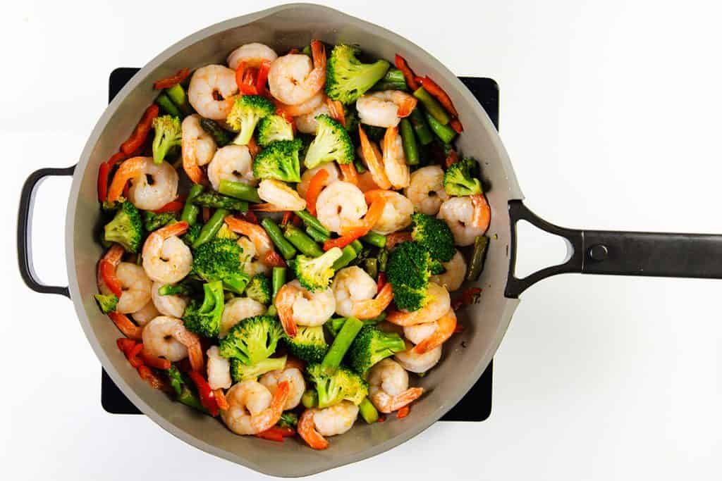 A skillet filled with cooked shrimp, broccoli florets, green beans, and red bell pepper slices makes an easy shrimp stir that's vibrant and delicious, viewed from above on a white background.