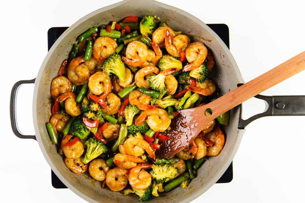 A pan of Shrimp Stir Fry with broccoli, snap peas, and red bell peppers is tossed in a glossy sauce. A wooden spatula rests in the pan on the stove—an easy shrimp recipe perfect for a quick dinner against a white background.