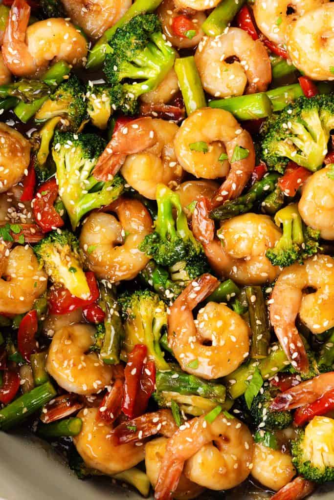 Close-up of a colorful shrimp stir-fry with broccoli, red bell pepper, and asparagus, coated in a glossy sauce and sprinkled with sesame seeds.
