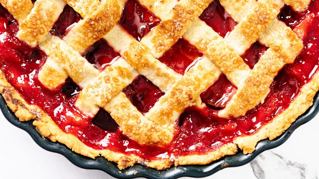 A close-up of a Homemade Strawberry Pie with a golden, lattice crust and glistening red filling in a black pie dish.