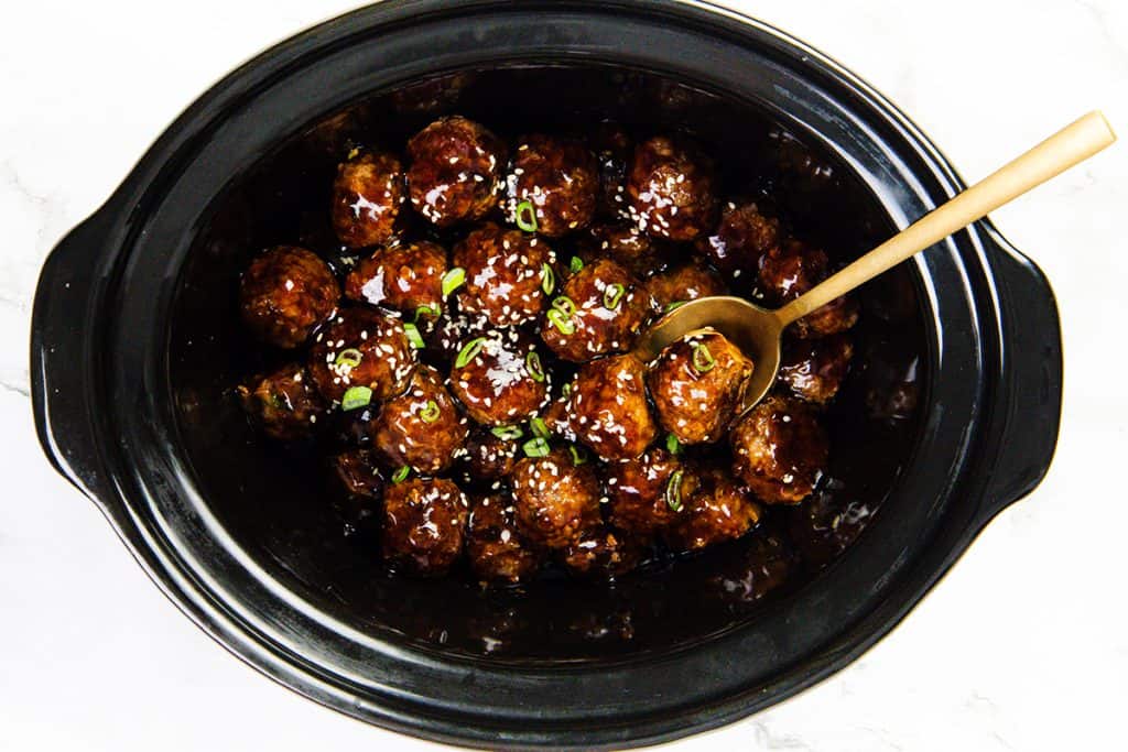 A black slow cooker filled with Teriyaki Meatballs glazed to perfection, garnished with chopped green onions and sesame seeds. A gold-colored serving spoon rests among the meatballs.