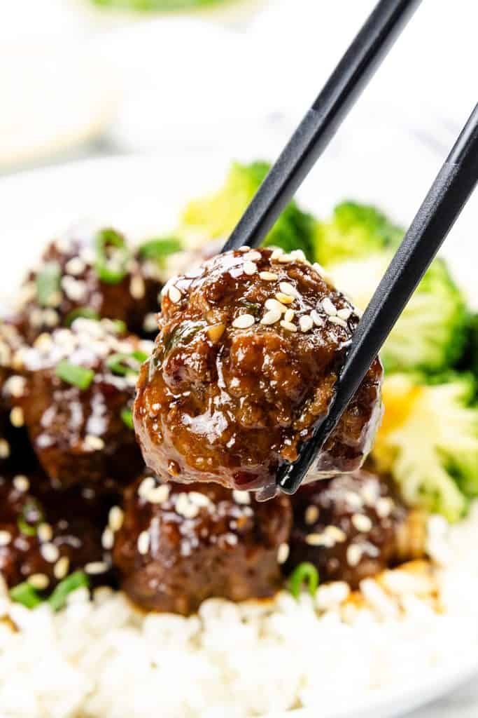Close-up of chopsticks holding a glazed Teriyaki Meatball sprinkled with sesame seeds above a plate of rice, broccoli, and more meatballs, all garnished with sesame seeds and green onions.