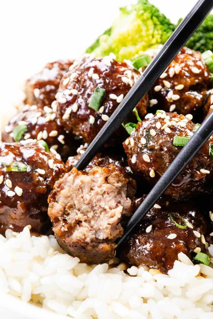 Chopsticks hold a glazed Teriyaki Meatball over white rice, surrounded by more meatballs topped with sesame seeds and chopped green onions, with broccoli in the background.