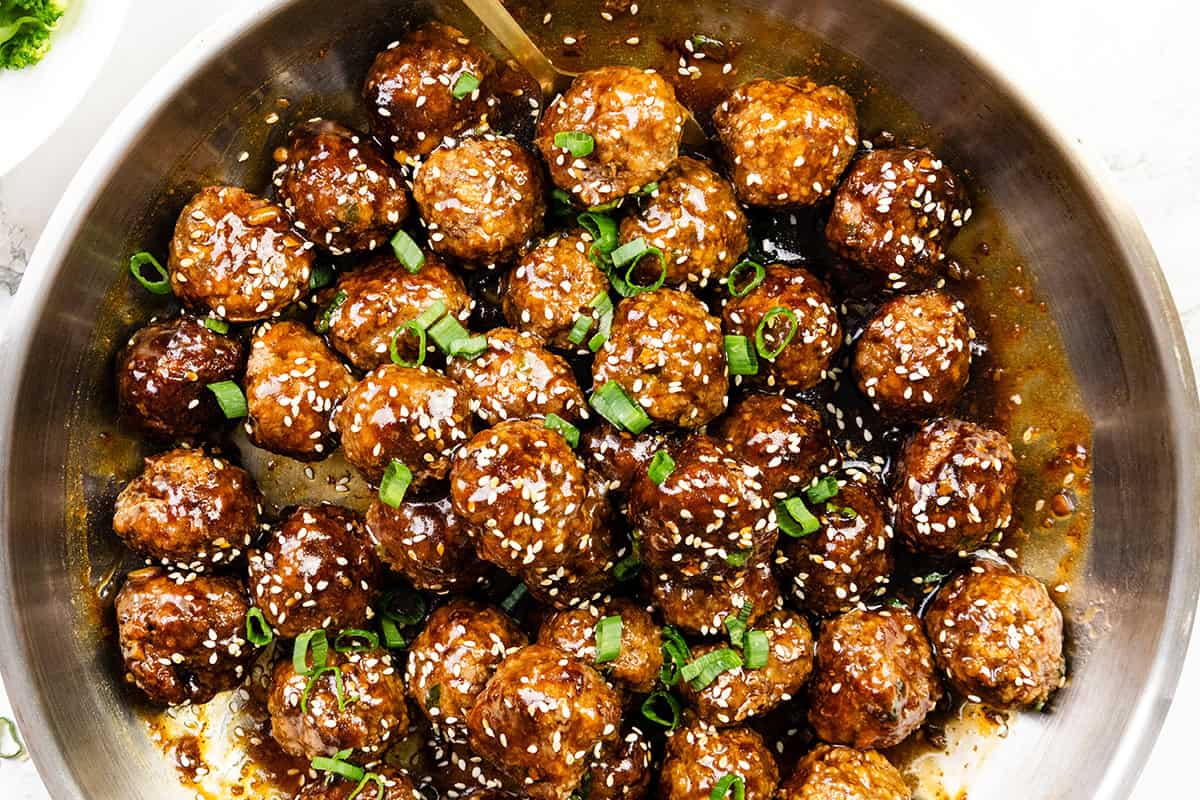 Teriyaki Meatballs coated in a glossy brown sauce, topped with sesame seeds and chopped green onions, served in a metal pan.