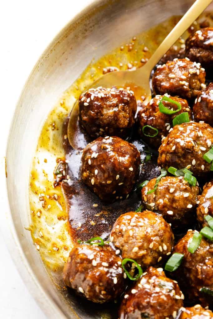 A skillet filled with Teriyaki Meatballs, glazed to perfection and topped with sesame seeds and sliced green onions, with a golden spoon resting among them.