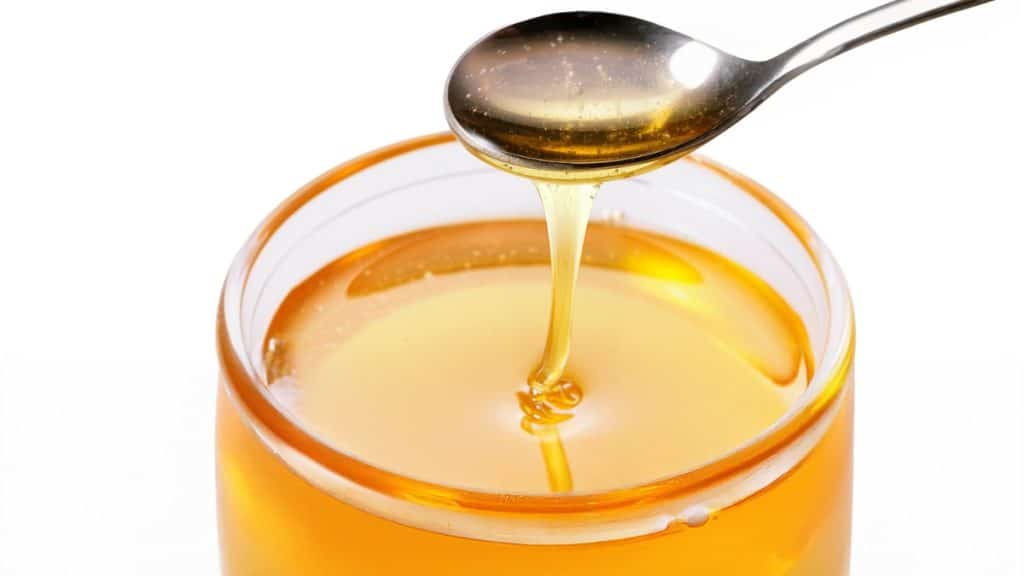 A spoon drizzling golden syrup into a clear glass jar filled with golden syrup, set against a white background—perfect for adding sweetness to your favorite treats like pecan pie.