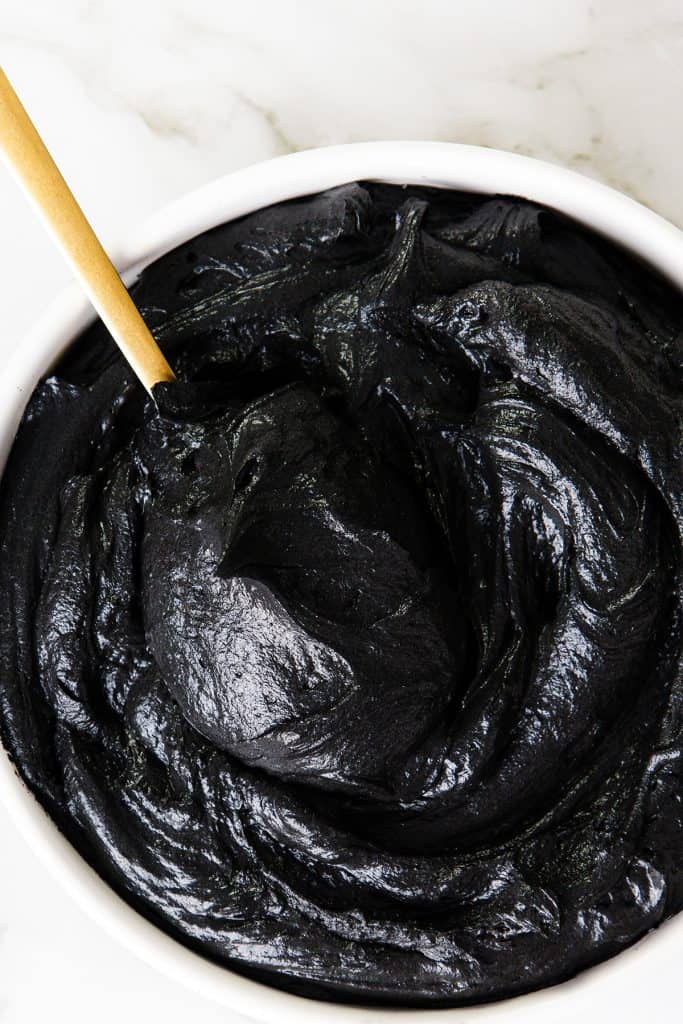 A white bowl filled with glossy black frosting being stirred with a gold-colored spoon, set on a light marble surface.