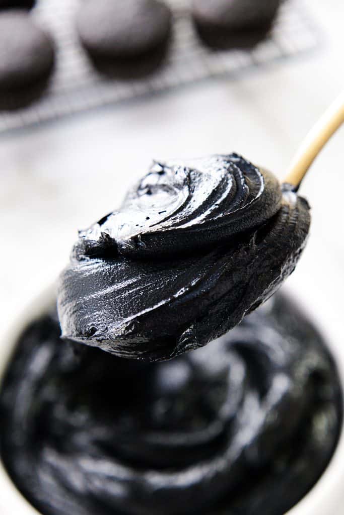 A close-up of a spoonful of glossy black frosting held above a bowl filled with more black frosting; blurred cookies on a wire rack add depth to this Black Frosting scene.