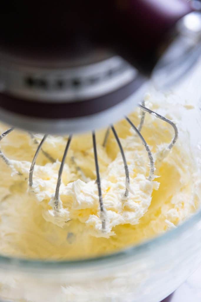 Close-up of a stand mixer with a wire whisk attachment, whipping a creamy, light yellow mixture in a glass bowl—perfect for creating a smooth base before adding bold black frosting accents.