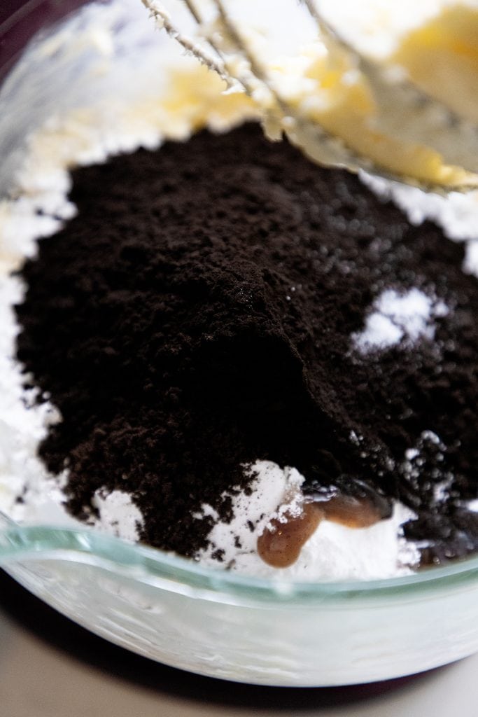 A close-up of a glass mixing bowl containing baking ingredients for Black Frosting, including dark cocoa powder, powdered sugar, and what appears to be softened butter and vanilla extract. Mixer beaters are visible at the top.