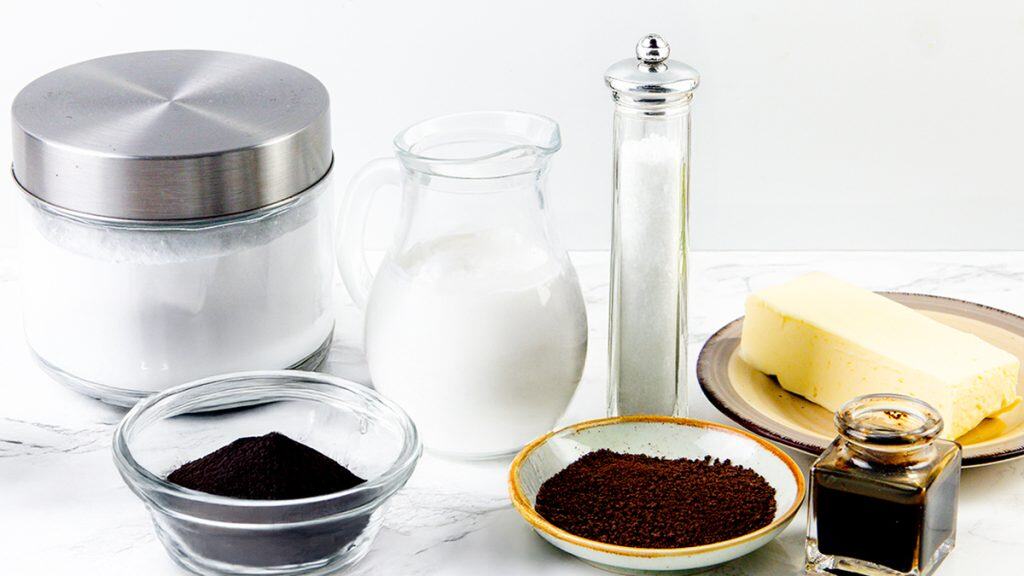 A collection of baking ingredients on a white surface, featuring a jar of flour, a pitcher of milk, butter on a plate, a tall salt shaker, sugar, vanilla, cocoa, coffee, and the essential supplies for making rich black frosting.
