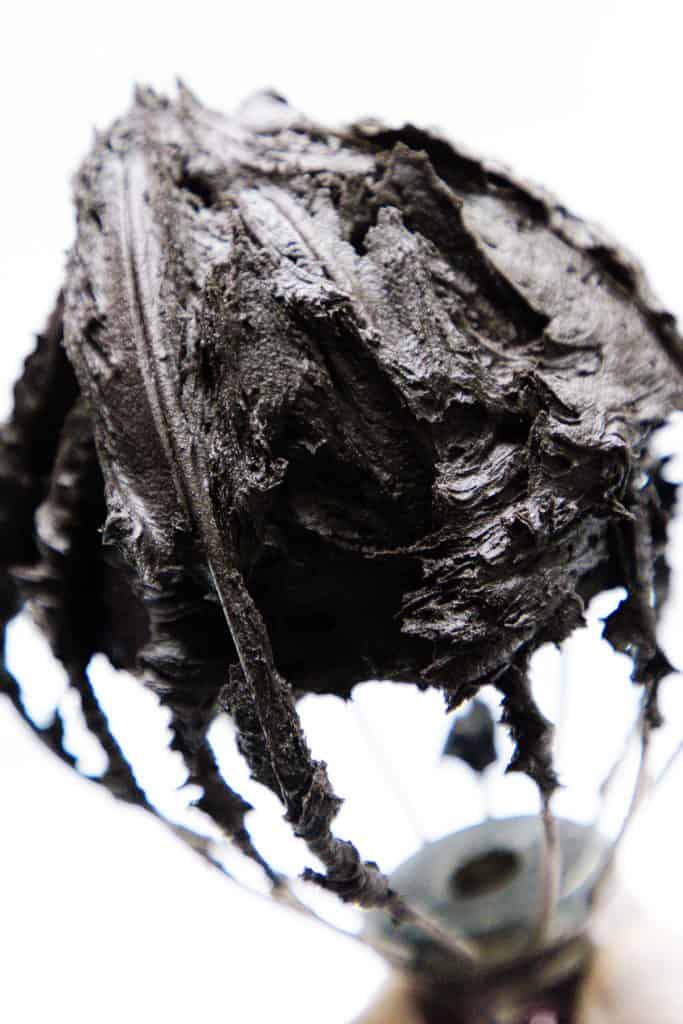 A close-up of a metal whisk covered in thick, black frosting and dark chocolate frosting against a white background.