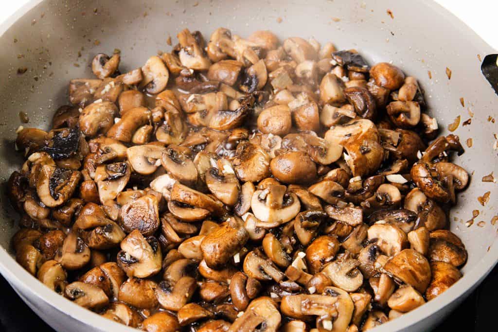 Sliced brown mushrooms being sautéed in a pan, releasing steam and mingling with bits of garlic and onion—the perfect start for a Creamy Mushroom Casserole.
