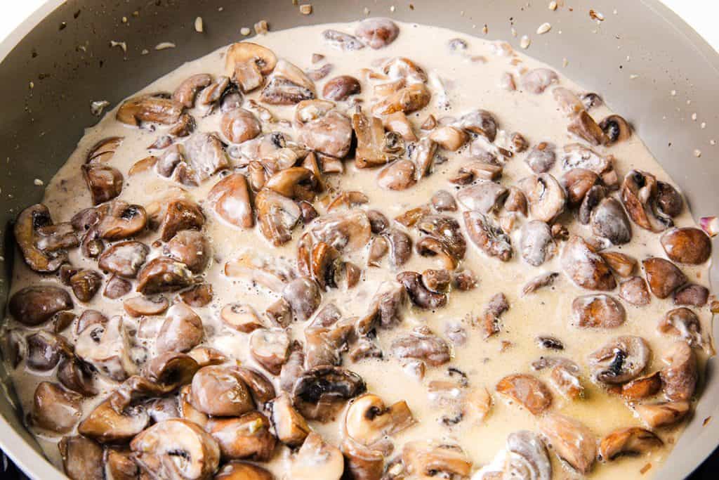 Sliced mushrooms cooking in a creamy sauce inside a large skillet, perfect for starting a delicious creamy mushroom casserole, with the mushrooms partially submerged in the light-colored sauce.