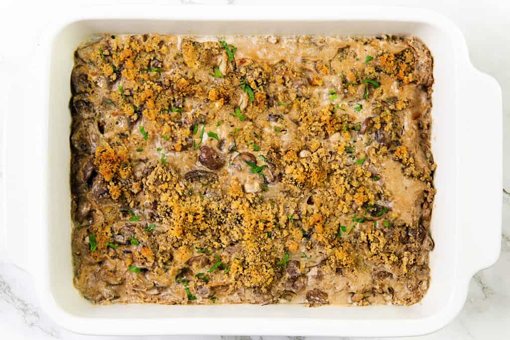 A white baking dish filled with creamy mushroom casserole, topped with golden brown breadcrumbs and garnished with chopped green herbs.