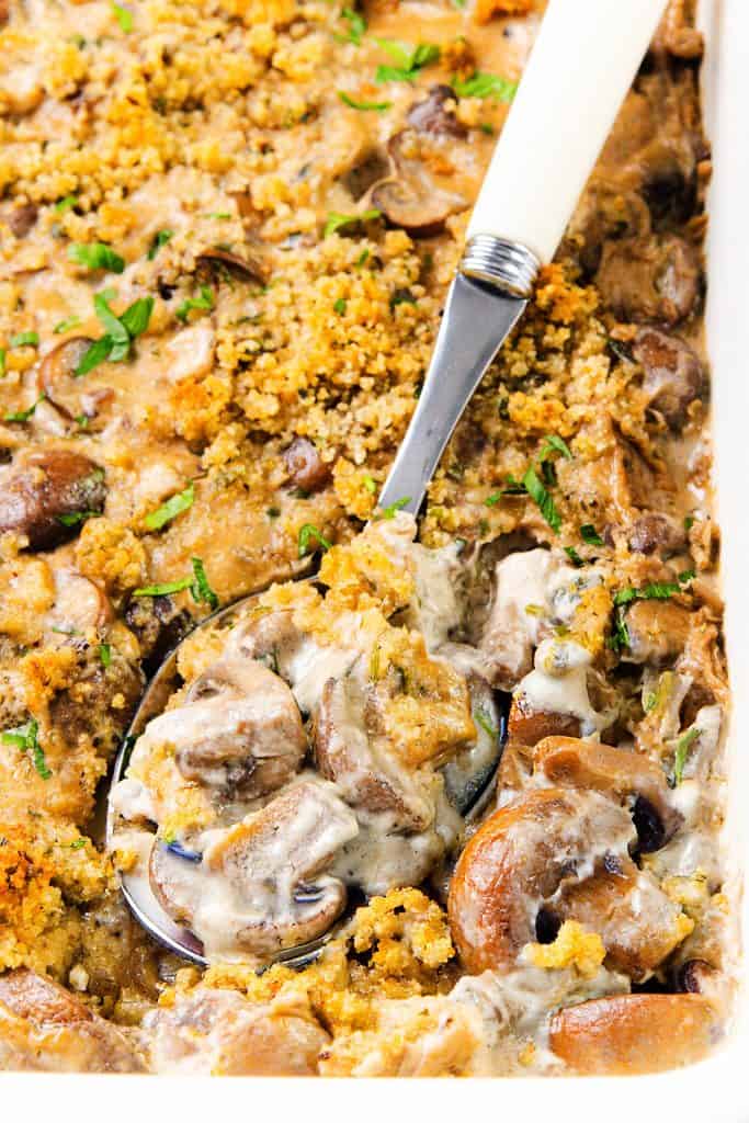 A close-up of a Creamy Mushroom Casserole topped with golden breadcrumbs and fresh herbs. A serving spoon lifts out a portion, revealing the rich, saucy texture and whole mushrooms inside this comforting casserole.