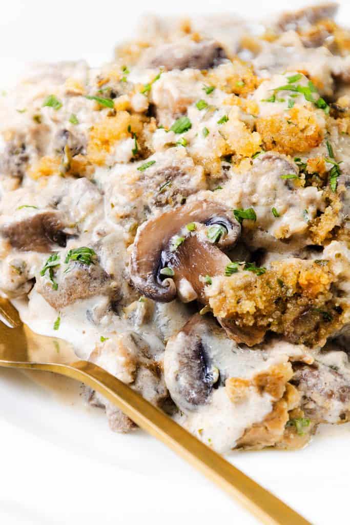 A close-up of Creamy Mushroom Casserole topped with golden breadcrumbs and fresh herbs, served with a gold-colored fork on a white plate.