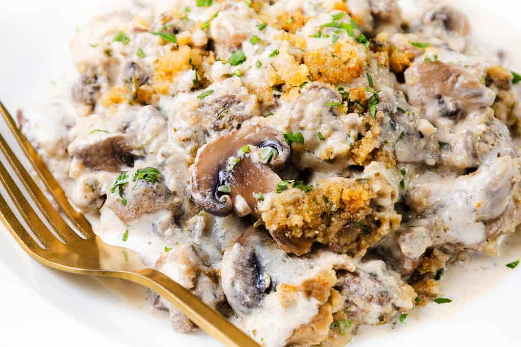 A close-up of a Creamy Mushroom Casserole with tender beef, golden breadcrumbs, and fresh herbs, served on a white plate with a gold fork.