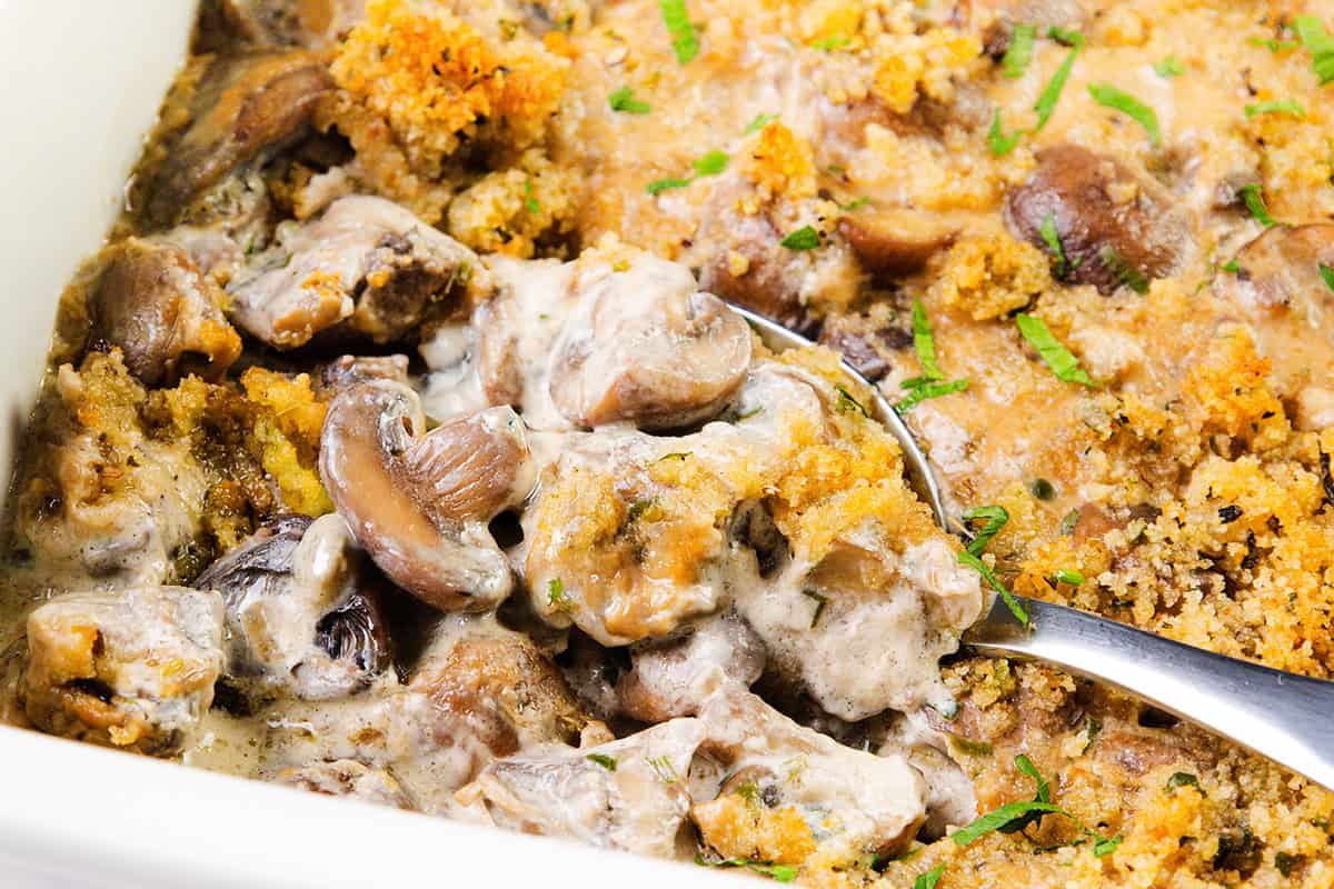 A close-up of creamy mushroom casserole topped with golden breadcrumbs and chopped herbs, with a spoon scooping out a portion, highlights the rich texture of this comforting Creamy Mushroom Casserole.