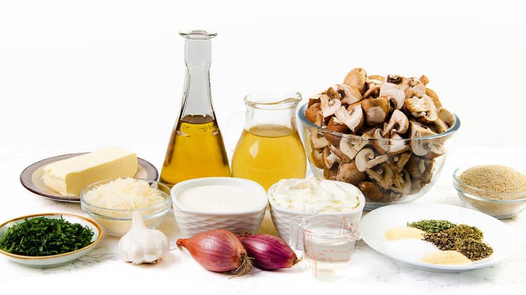 A variety of ingredients on a white surface for a Creamy Mushroom Casserole, including chopped mushrooms in a glass bowl, blocks of butter, grated cheese, fresh herbs, garlic, shallots, olive oil, broth, cream, breadcrumbs, and assorted seasonings.