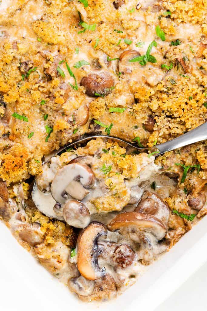 A close-up of a Creamy Mushroom Casserole topped with golden breadcrumbs and fresh herbs, with a spoon scooping out a hearty portion.