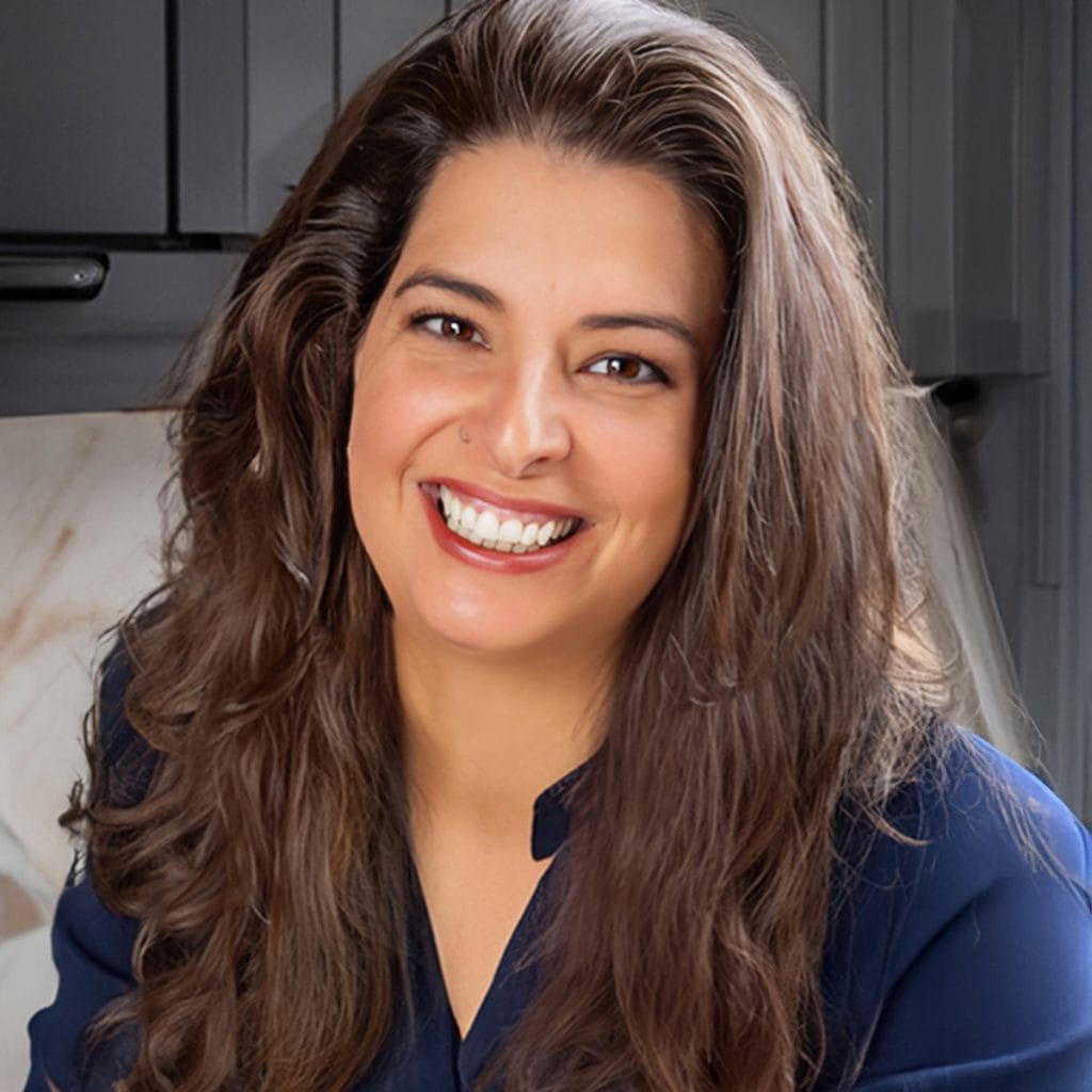 About Erren Hart: A woman with long brown hair and a wide smile, wearing a dark blue blouse, sits in a modern kitchen with dark cabinets and a marble backsplash.