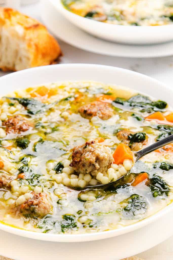 A close-up of Italian Wedding Soup featuring meatballs, small pasta, spinach, and diced carrots in broth. A spoon inside the bowl holds a meatball, pasta, and vegetables. A piece of bread is visible in the background.