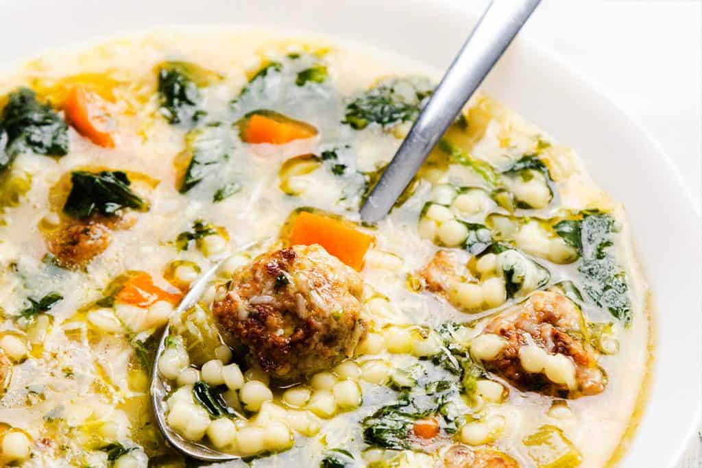 A close-up of Italian Wedding Soup in a white bowl, featuring savory meatballs, carrots, leafy greens, and small pasta, with a spoon scooping up some of the ingredients.