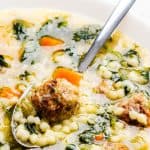 A close-up of Italian Wedding Soup in a white bowl, featuring savory meatballs, carrots, leafy greens, and small pasta, with a spoon scooping up some of the ingredients.