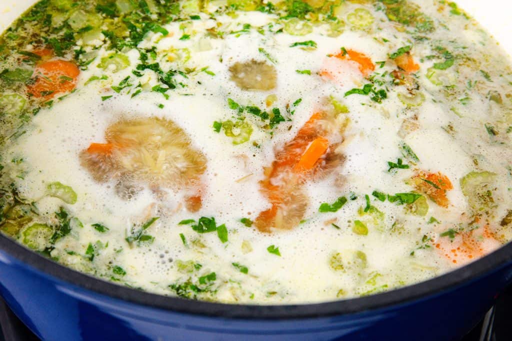 A close-up of a simmering pot of one-pot soup with visible chunks of carrots, green herbs, and meat pieces in a blue pot. The broth is bubbling gently, with foam on the surface for a cozy homemade feel.