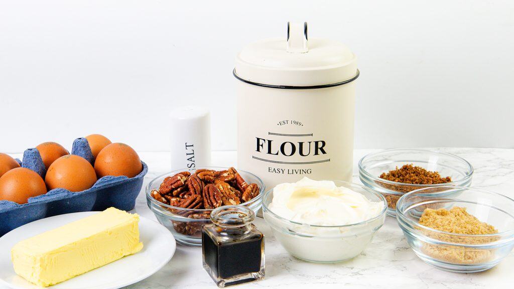 A collection of baking ingredients for delicious treats like pecan cookies or Pecan Tassies, including eggs, butter, pecans, vanilla extract, salt, flour, brown sugar, and cream cheese, arranged neatly on a white countertop.