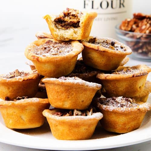 A stack of Mini Pecan Pies arranged on a white plate, with a bowl of pecans and a flour container in the background on a white surface.
