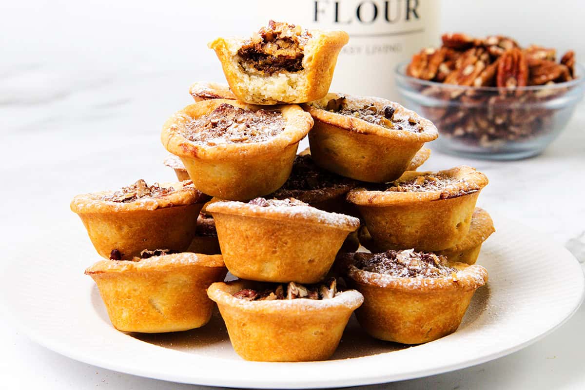 A stack of Mini Pecan Pies arranged on a white plate, with a bowl of pecans and a flour container in the background on a white surface.