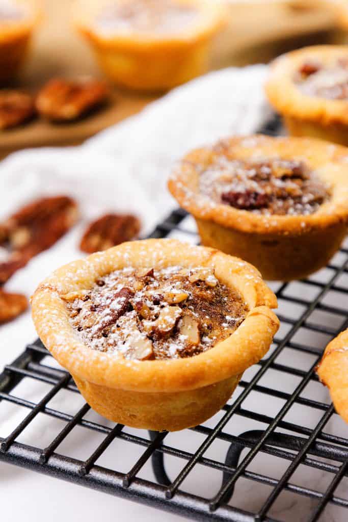 Mini Pecan Tassies topped with powdered sugar cool on a black wire rack, surrounded by whole pecans and a soft white cloth in the background.