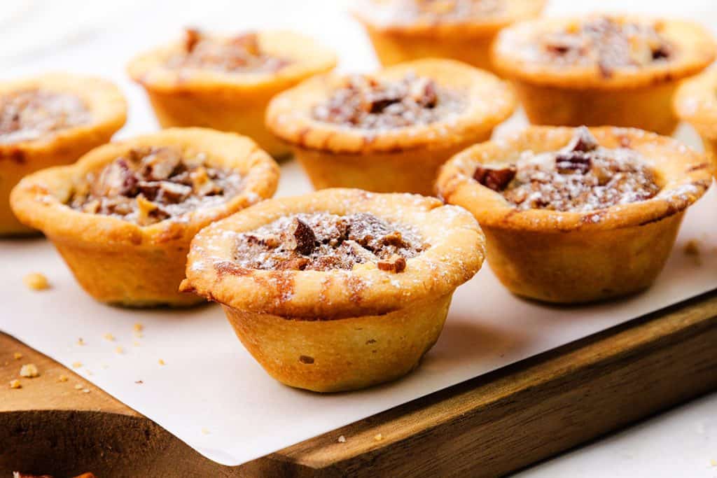 A close-up of several Pecan Tassies on a parchment-lined wooden board. The mini pecan pies have golden-brown crusts and are filled with chopped pecans, with a few crumbs scattered around them.