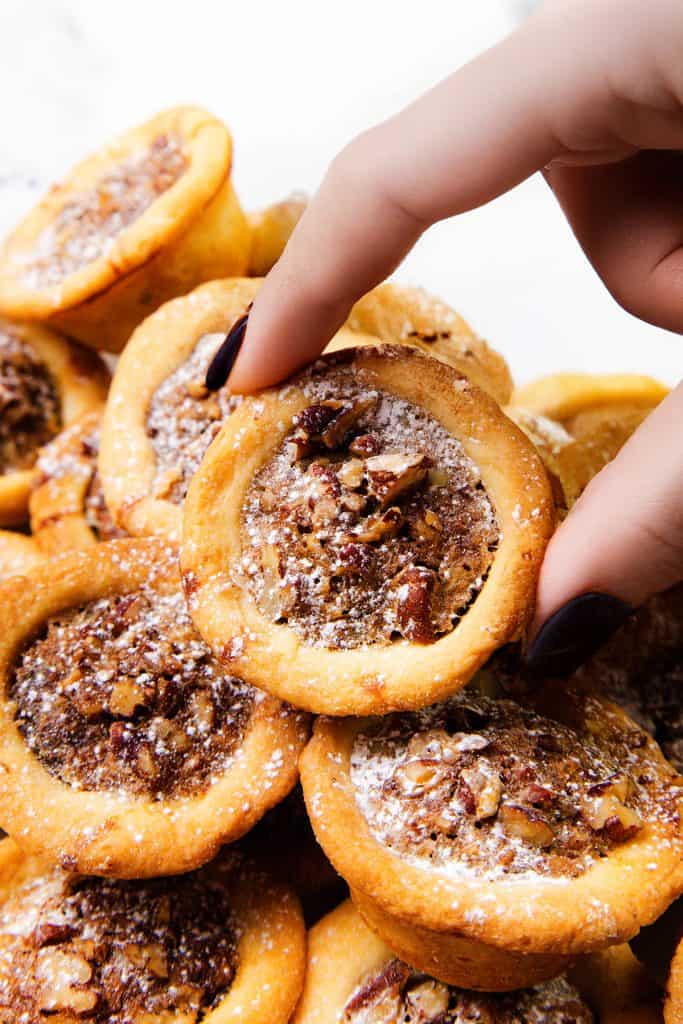 A hand with dark nail polish lifts a mini pecan pie dusted with powdered sugar from a pile of similar pecan desserts.