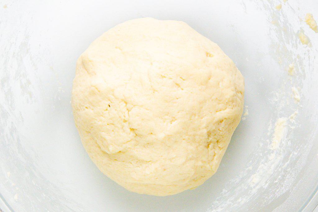 A ball of pale, smooth dough sits in a clear glass bowl, ready to be transformed into delicious Mini Pecan Pies. The dough has a slightly rough texture, and bits of flour cling to the sides of the bowl.