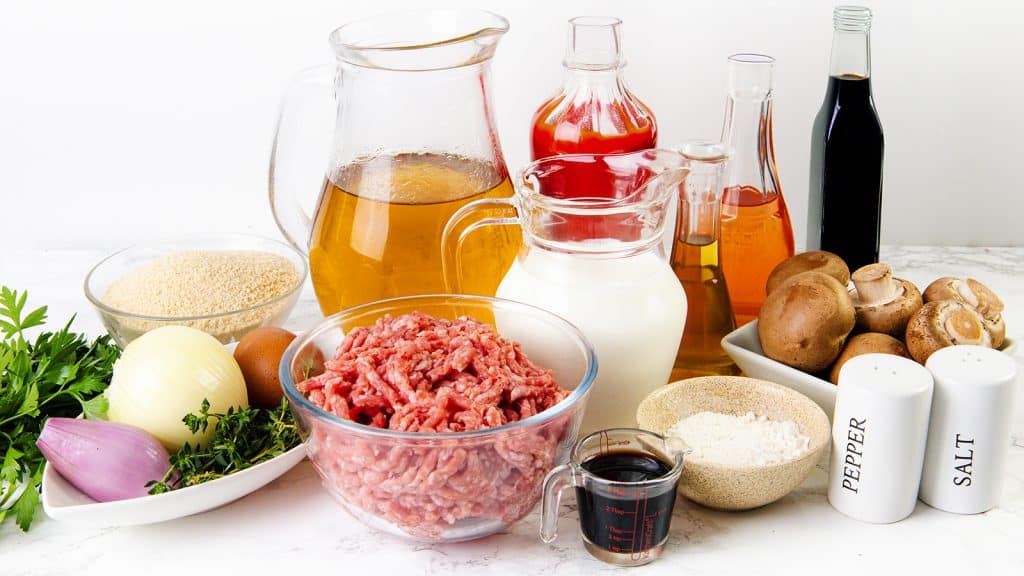 A variety of ingredients for Salisbury Steak on a white surface, including ground meat, eggs, mushrooms, onion, shallot, fresh herbs, couscous, milk, broth, vinegars, Worcestershire sauce, flour, and salt and pepper shakers.
