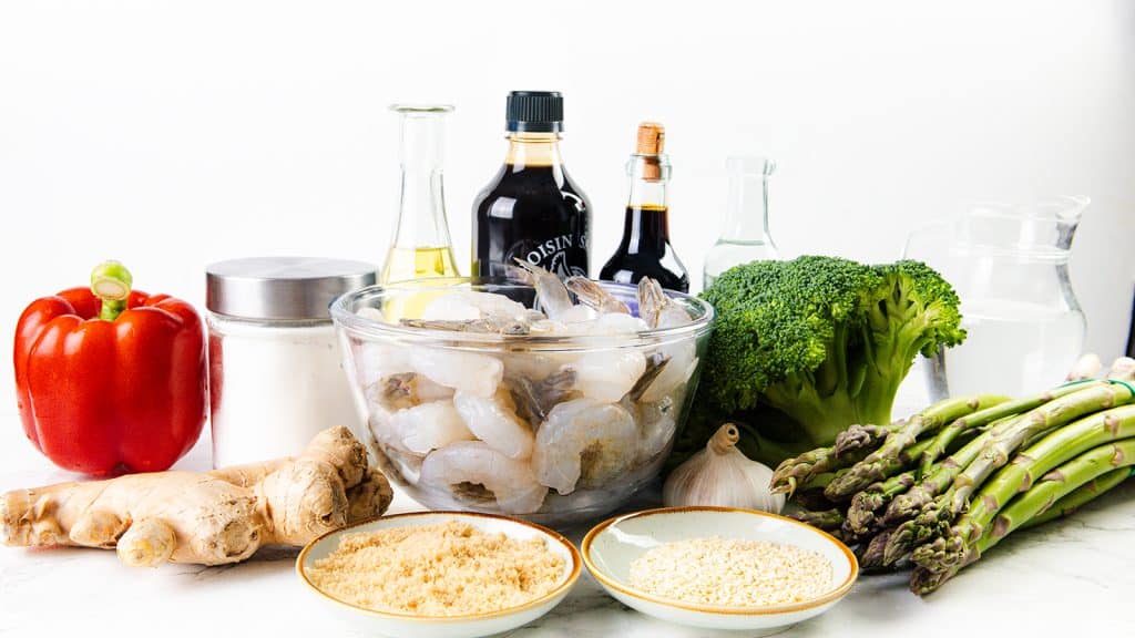 A variety of fresh and packaged ingredients for cooking shrimp stir-fry, including raw shrimp in a glass bowl, red bell pepper, ginger, brown sugar, sesame seeds, garlic, asparagus, broccoli, oils, sauces, and a jar of sugar on a white surface.
