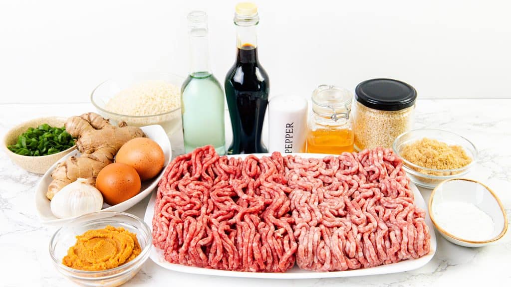 A platter of raw ground meat awaits transformation into Teriyaki Meatballs, surrounded by bowls of eggs, green onions, garlic, ginger, pumpkin puree, breadcrumbs, seasonings, soy sauce, vinegar, oil, and a jar of honey on a white countertop.