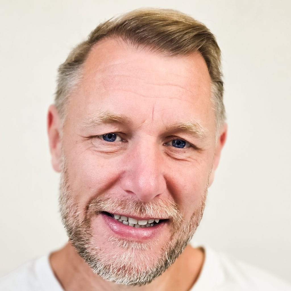 About Erren Hart: A middle-aged man with light skin, short blonde hair, and a neatly trimmed beard smiles at the camera against a plain light background. He is wearing a white shirt.